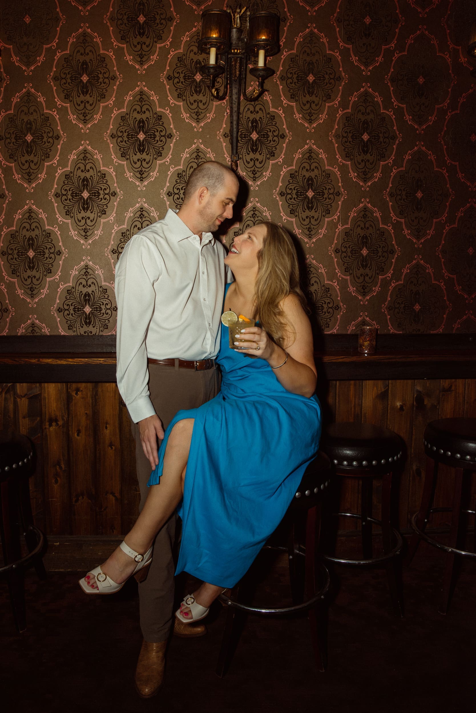 Woman in a blue dress on a bar stool gazing up at her fiancé in front of damask wallpaper and a candle sconce