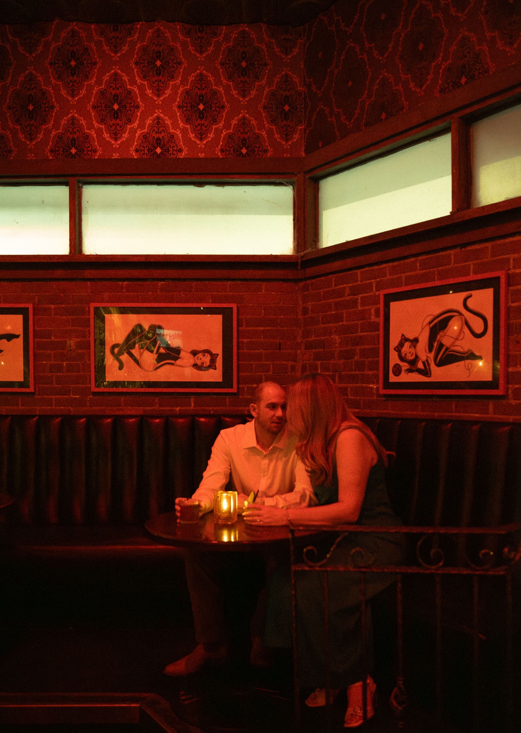 Couple sharing a quiet moment in a red-lit corner booth beneath cat pin-up prints and high windows