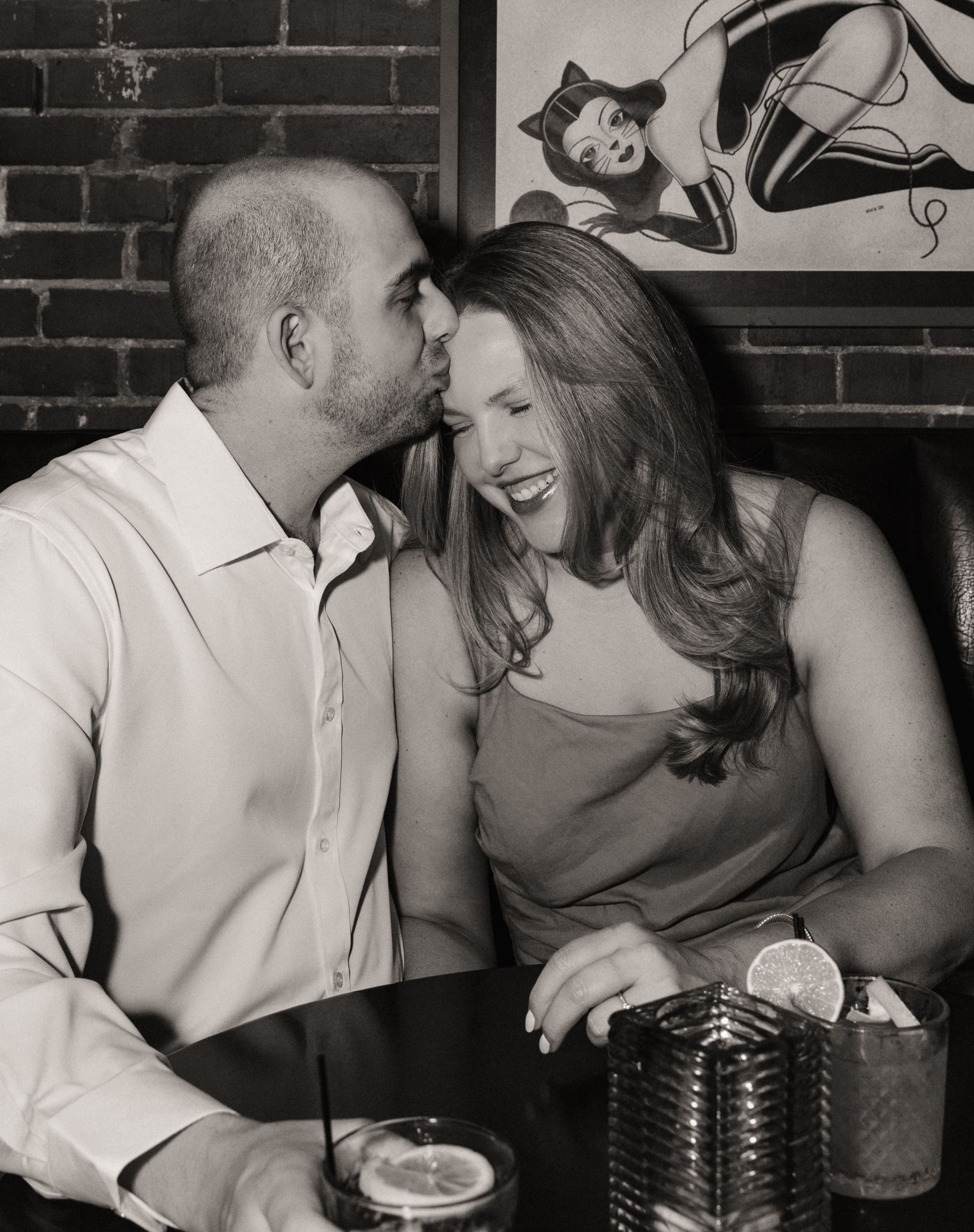 Black and white photo of a man kissing his laughing fiancée's temple in a booth beneath a pin-up print