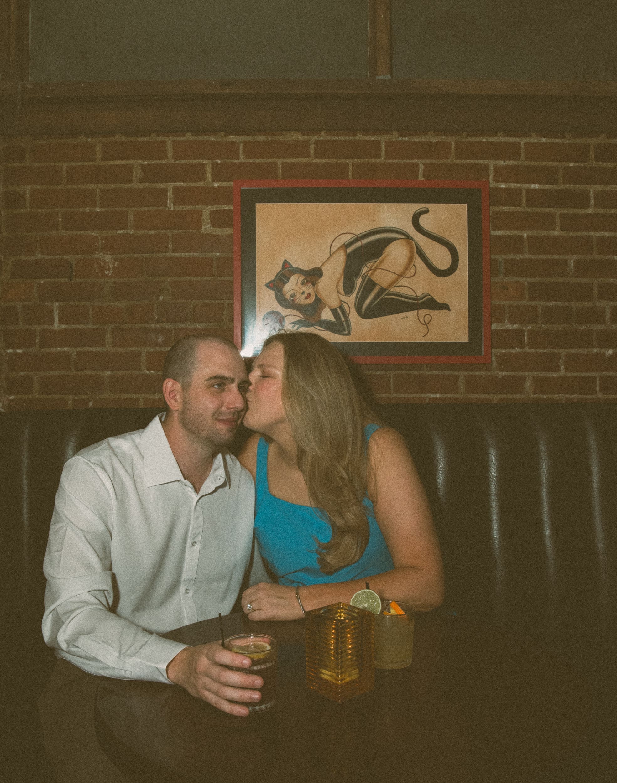 Woman kissing her fiancé's cheek in a leather booth beneath a cat pin-up print on exposed brick