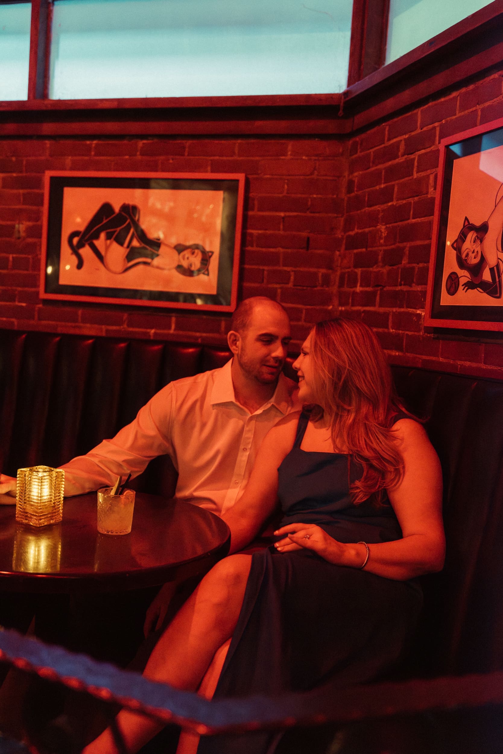 Engaged couple leaning close in a red-lit corner booth between two framed pin-up prints