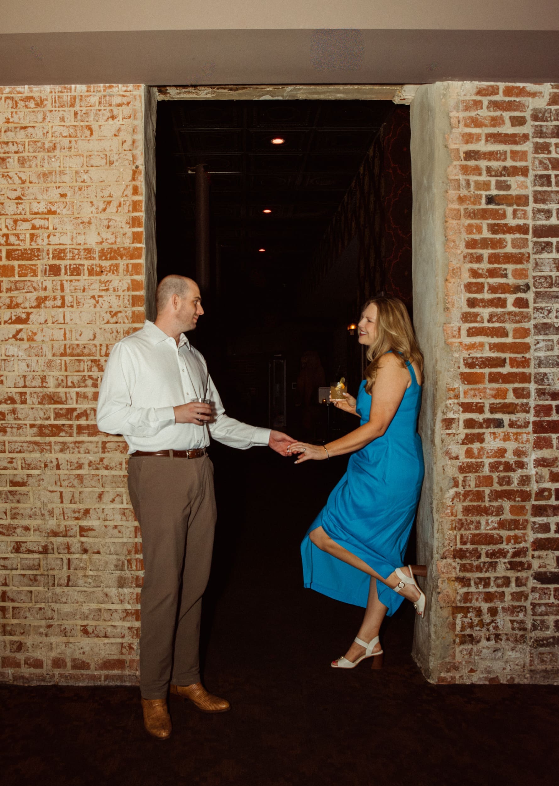 Couple holding hands in a brick doorway as she kicks one foot up against the wall, drink in hand