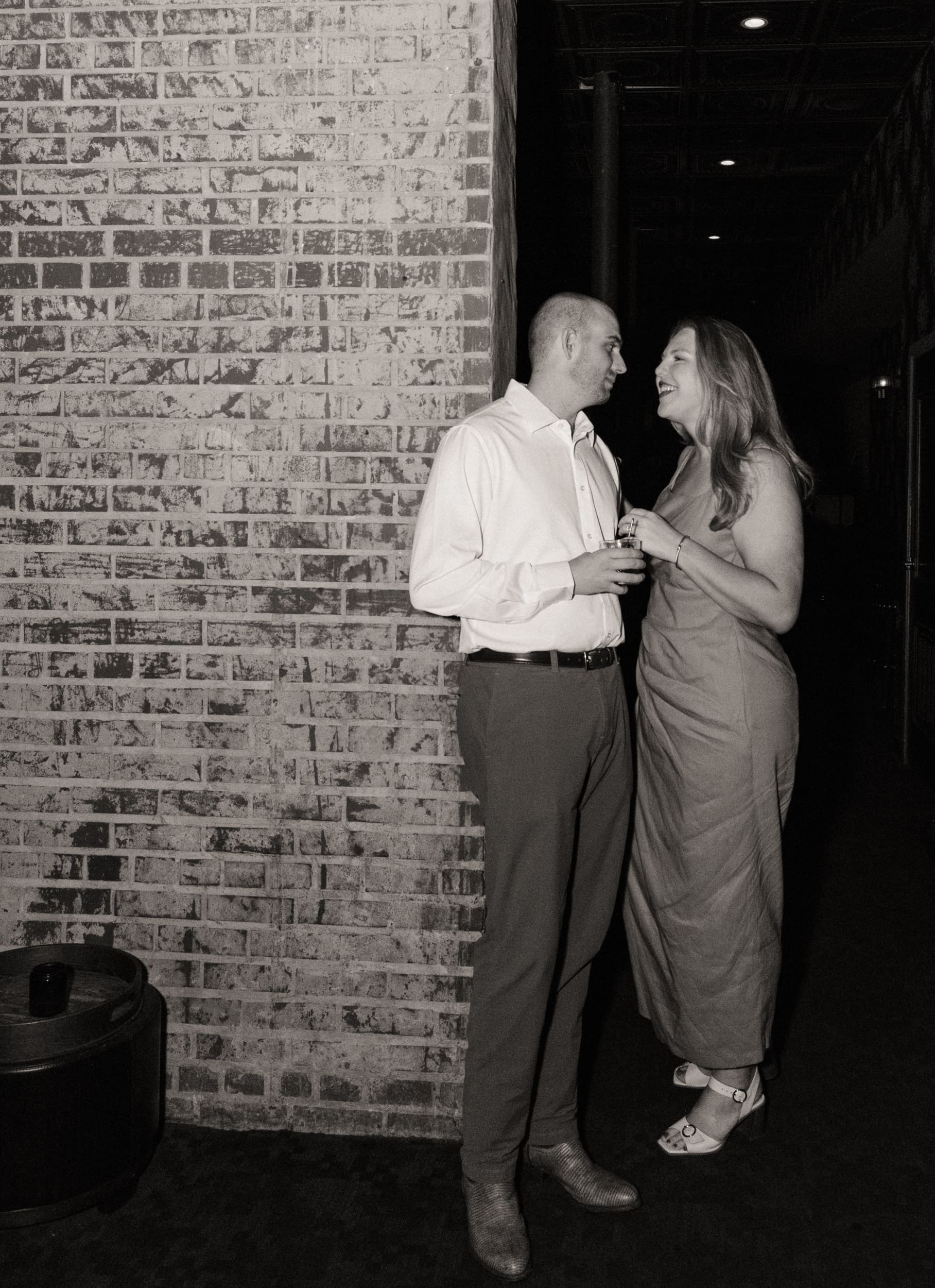 Black and white photo of an engaged couple standing close with cocktails in a brick doorway