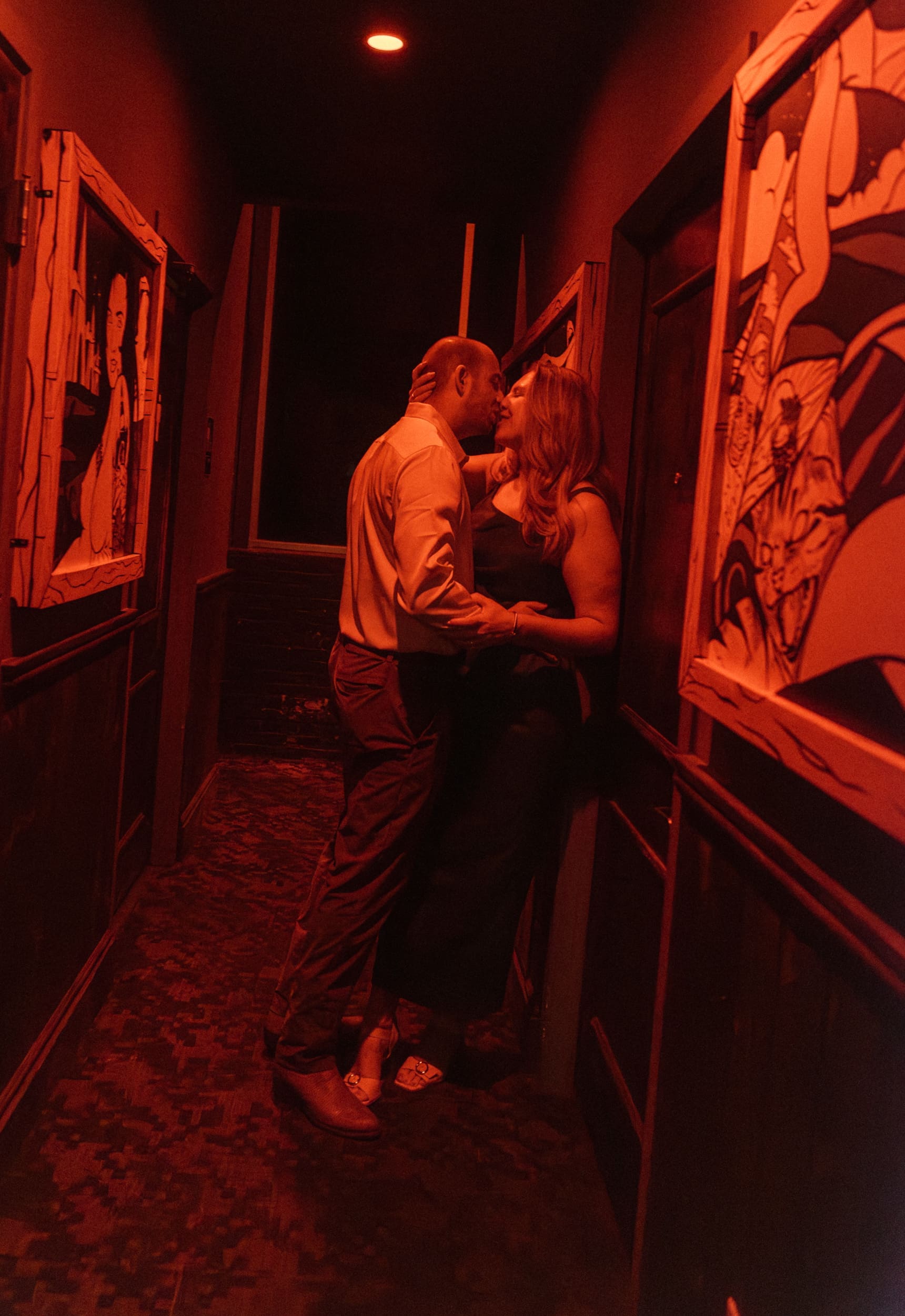 Couple kissing in a narrow red-lit hallway lined with bold comic-style murals at Dos Gatos