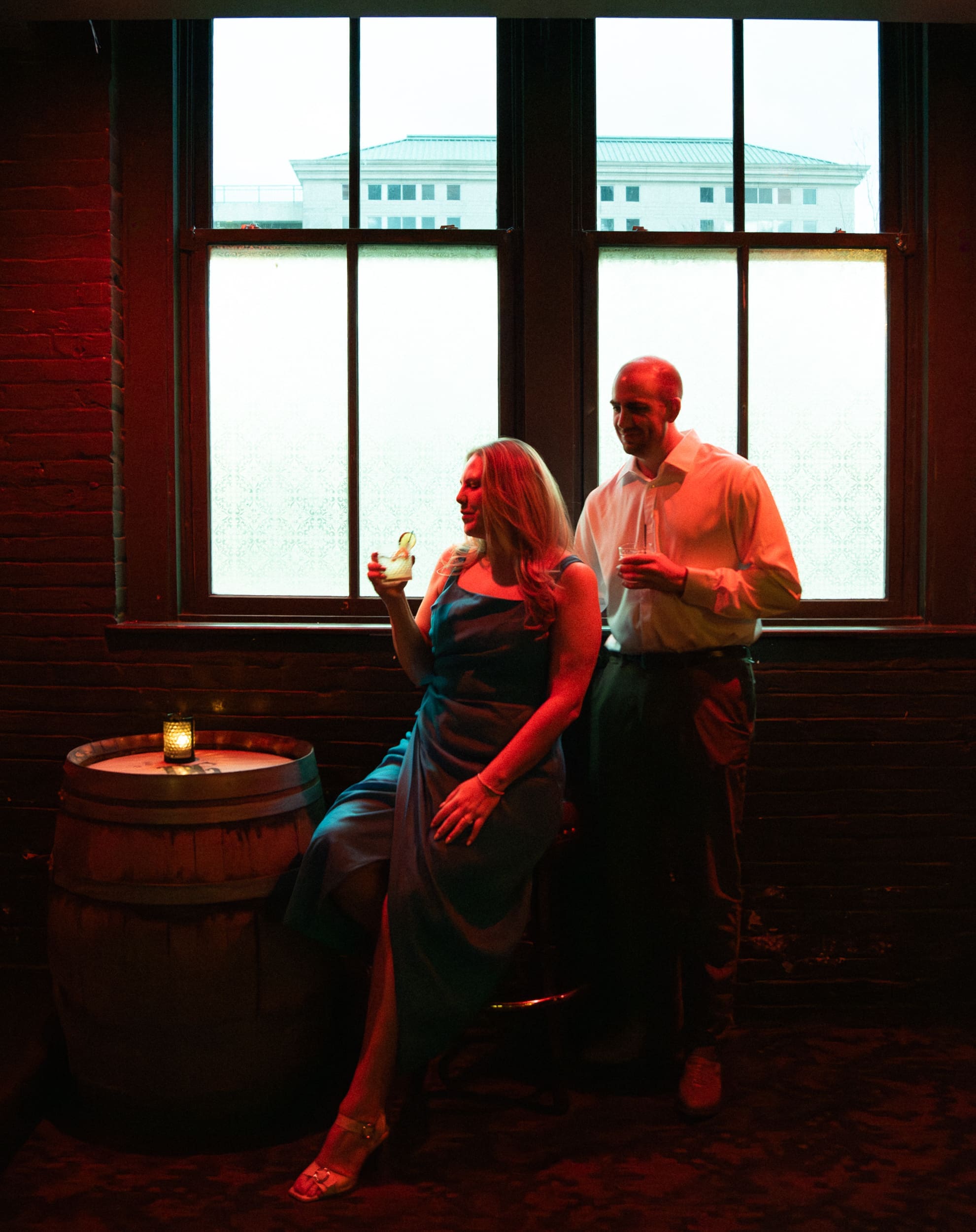 Engaged couple with cocktails in warm red light against a large frosted window at Dos Gatos