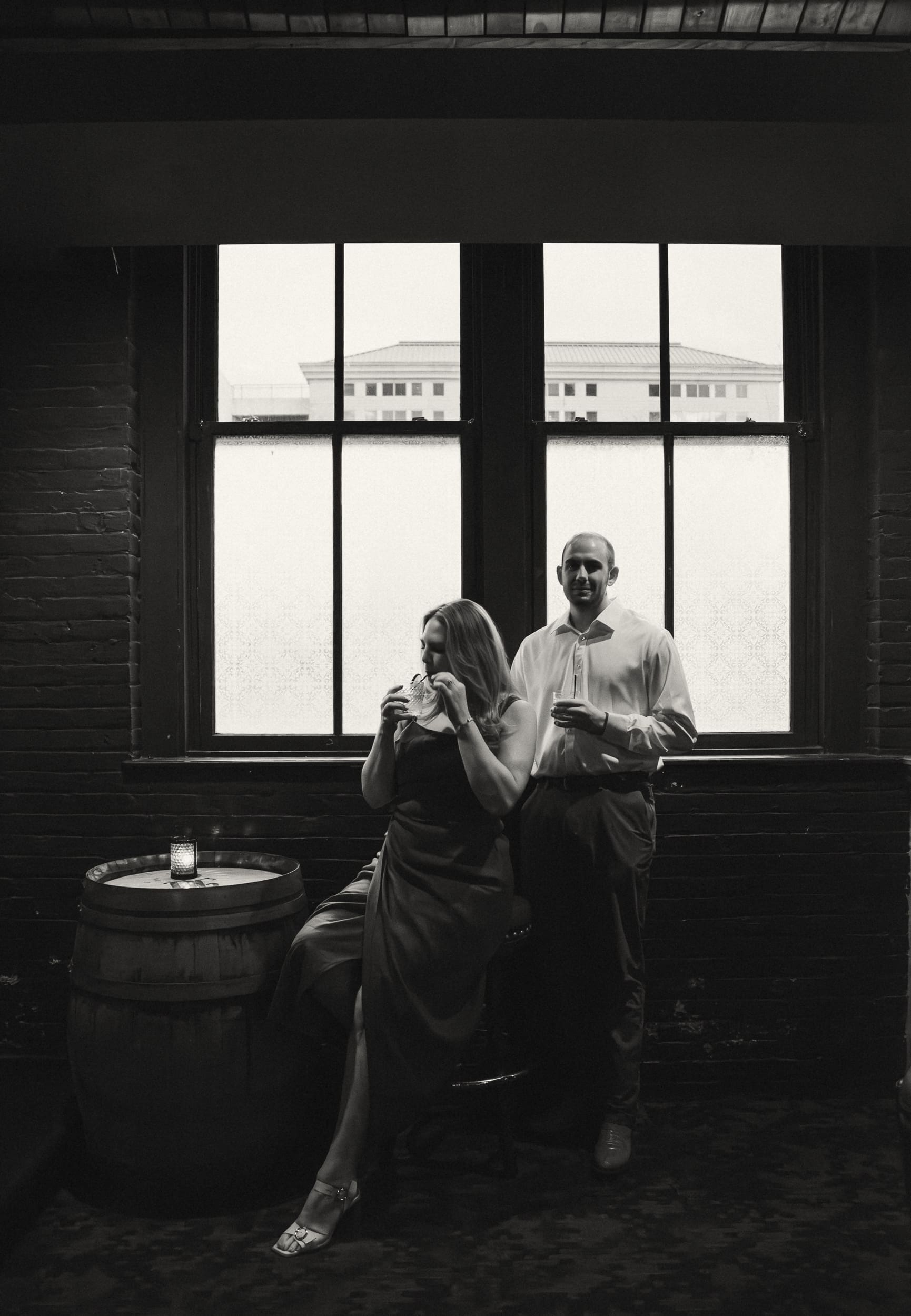 Black and white portrait of an engaged couple with drinks in front of a large window at Dos Gatos