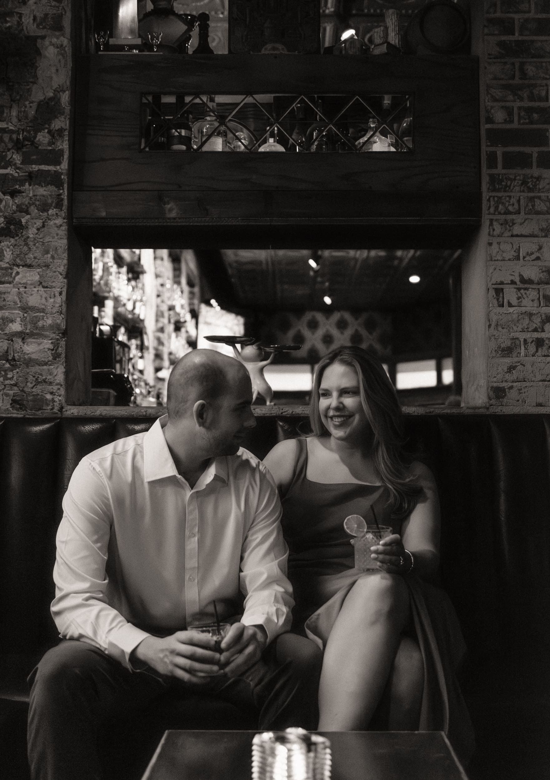 Black and white photo of a laughing couple with drinks in a booth beneath a window into the backbar