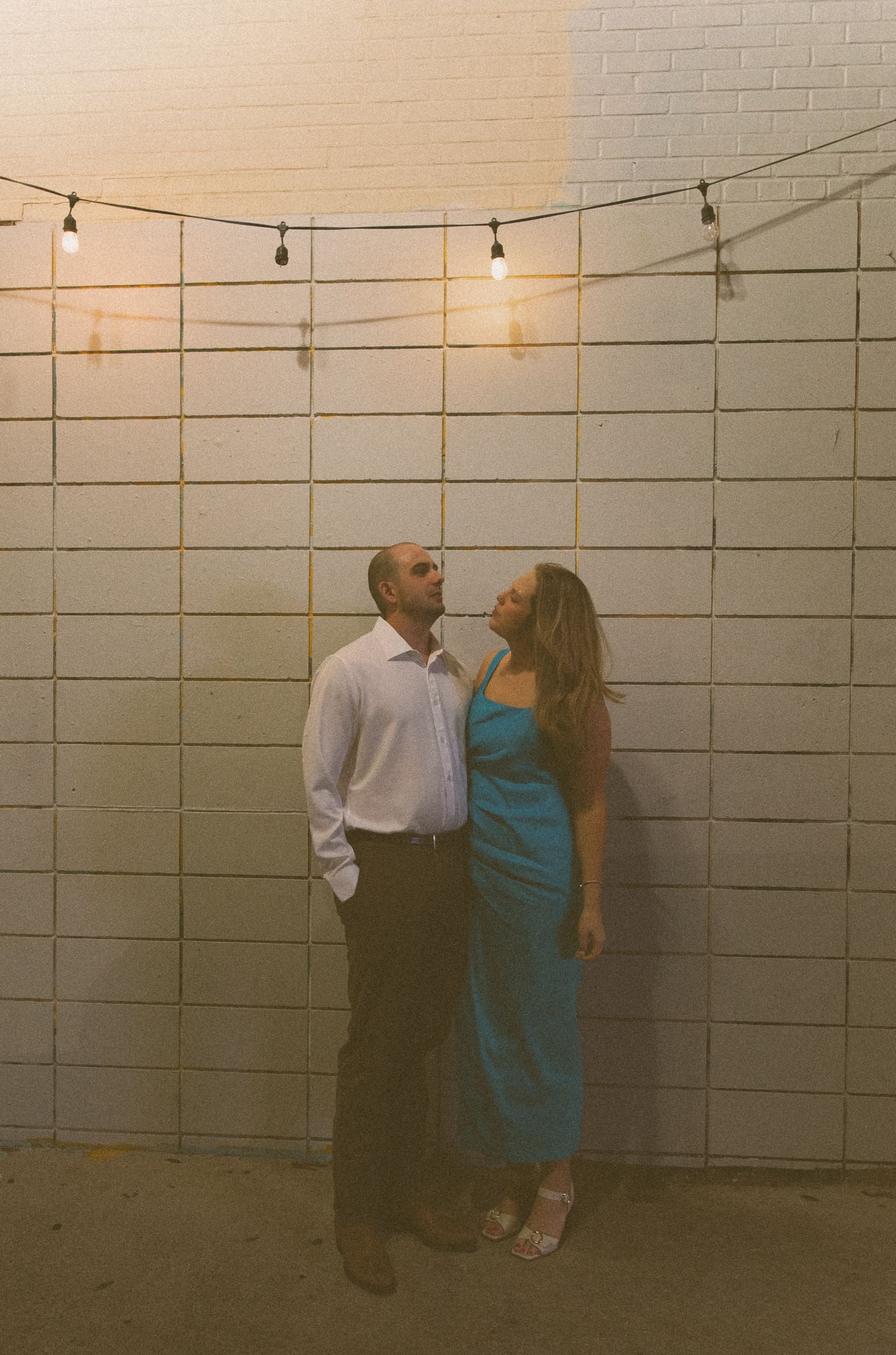 Couple standing close against a white brick wall beneath hanging string lights in downtown Jacksonville
