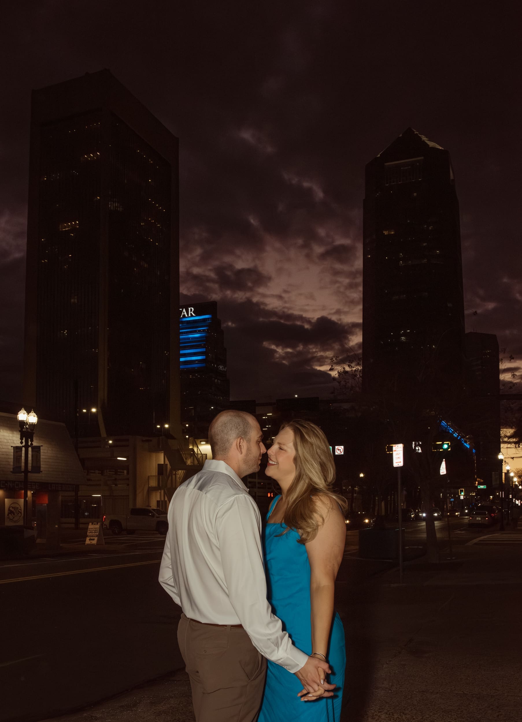 Engaged couple holding hands beneath a cloudy night sky framed by downtown Jacksonville skyscrapers